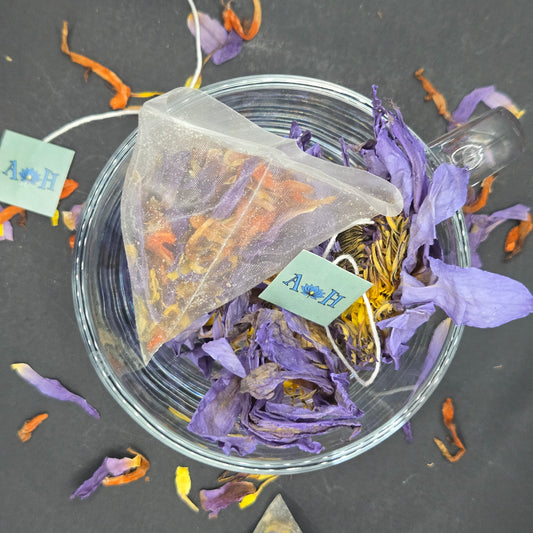 Astral Herbal tea bags with wild dagga, blue lotus, and chamomile in a glass teacup on a black background with loose blue lotus and wild dagga petals in the foreground.