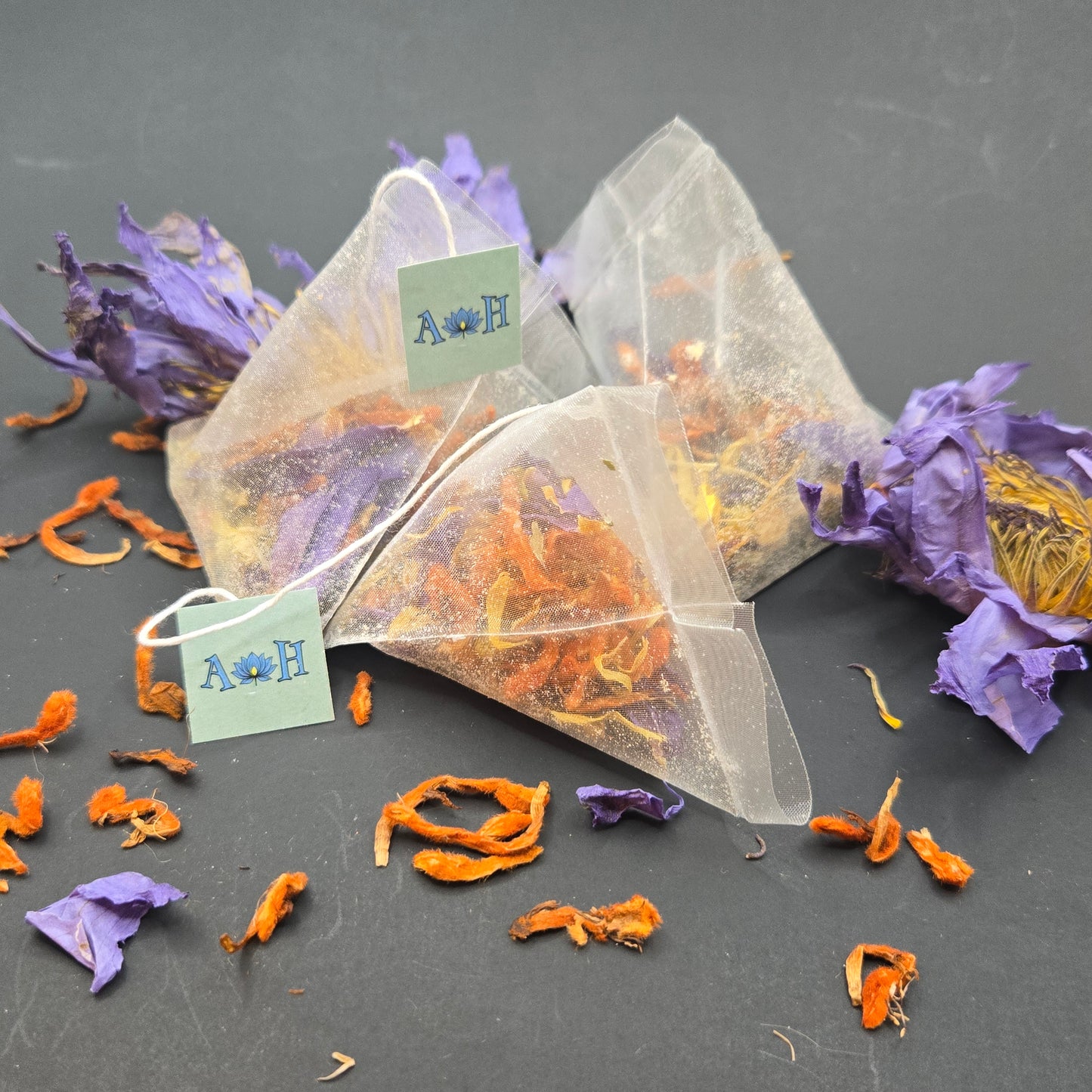 Astral Herbal pyramid tea bags with wild dagga, blue lotus, and chamomile on a black background with loose blue lotus and wild dagga petals in the foreground.