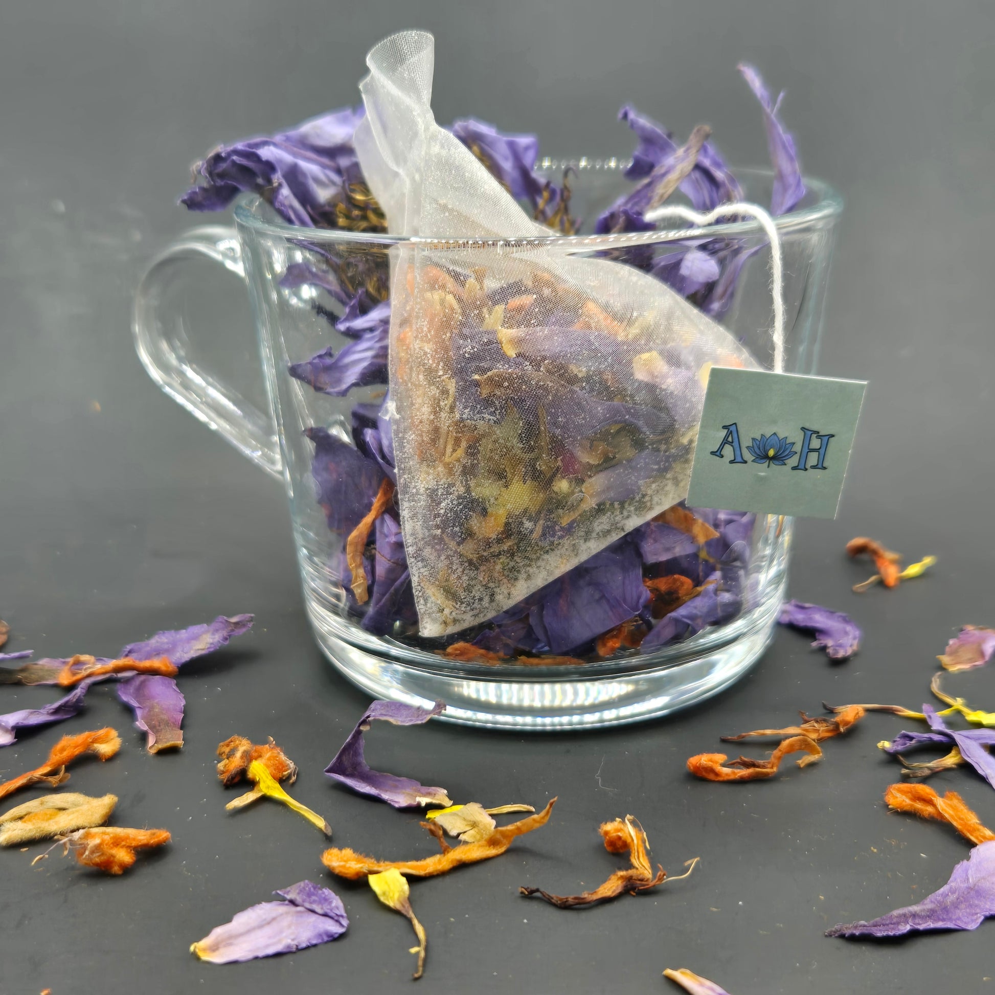 Astral Herbal tea bag with wild dagga, blue lotus, and chamomile in a glass teacup on a black background with loose blue lotus and wild dagga petals in the foreground.